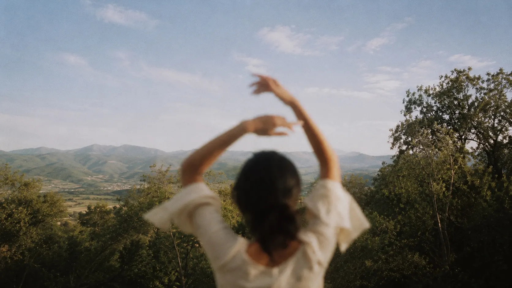 A woman seen from behind, arms raised overhead, dancing on a hilltop above green valleys