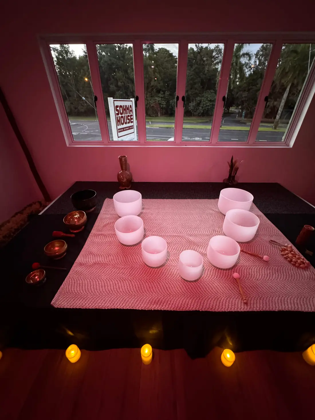 Crystal and brass singing bowls set out on a pink-lit cloth with candles along the floor, and the Sohma House sign standing in the window beyond.