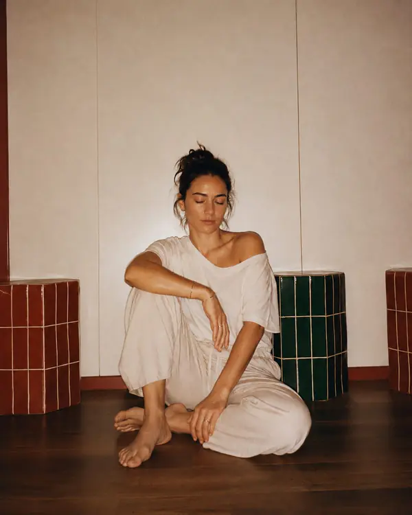 A woman sitting on a timber floor in loose linen, eyes closed, at ease.
