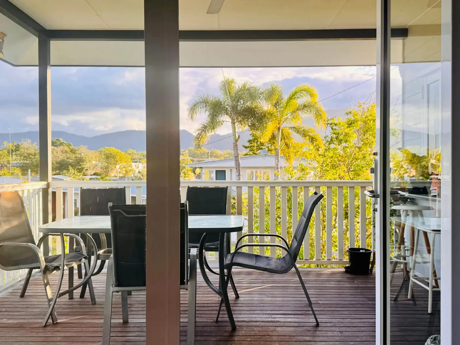 The upstairs verandah at golden hour: a table and chairs, a white rail, palms and the ranges beyond.